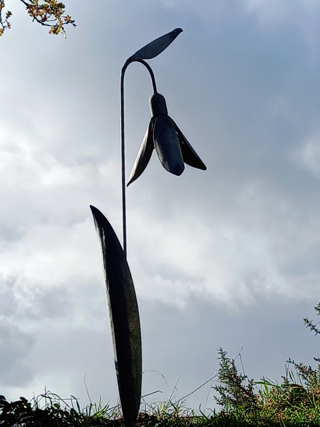 A tall metal sculpture of a snowdrop flower with a solid gray color, standing in an outdoor setting with a cloudy sky in the background.