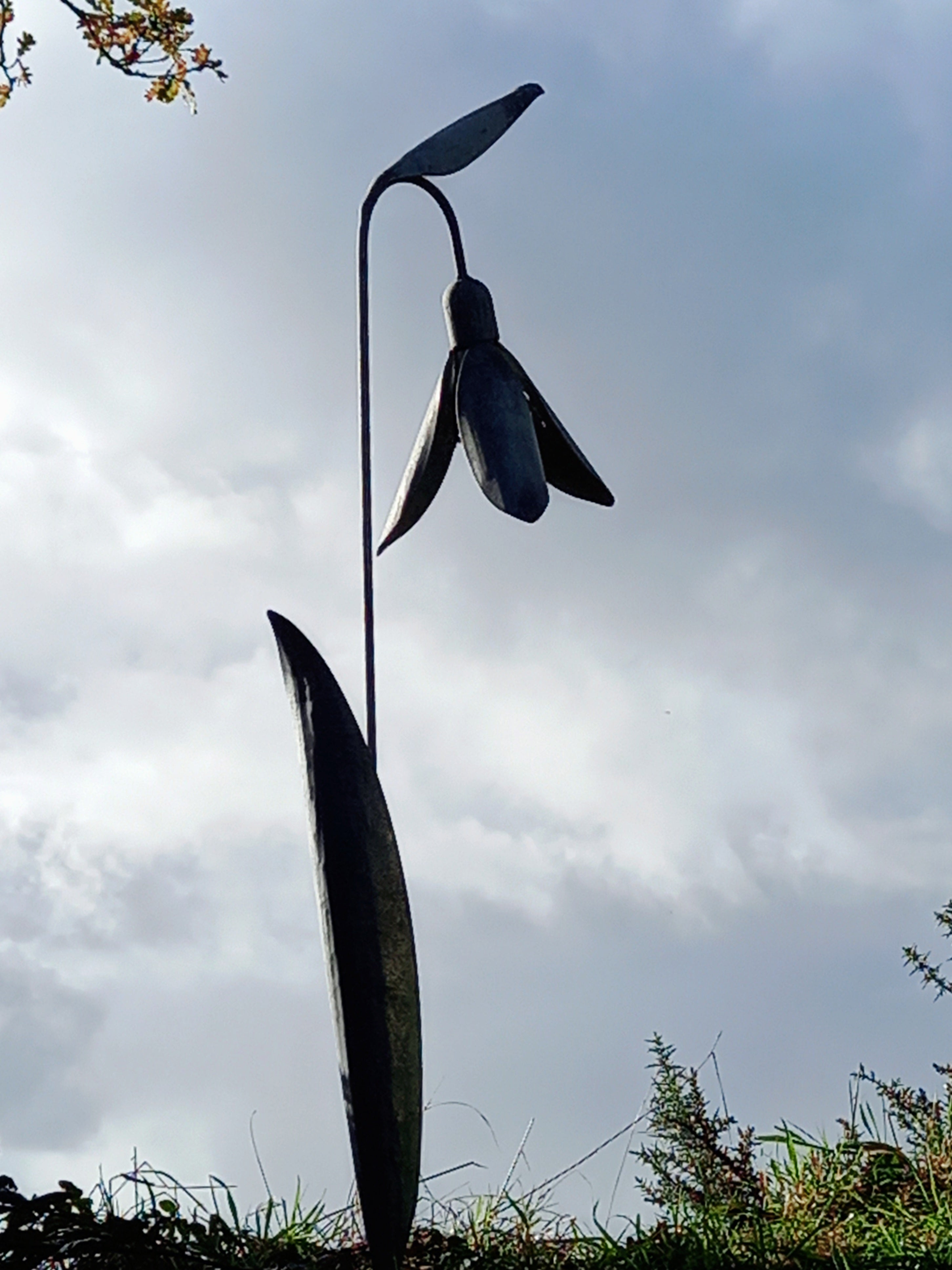 A tall metal sculpture of a snowdrop flower with a solid gray color, standing in an outdoor setting with a cloudy sky in the background.