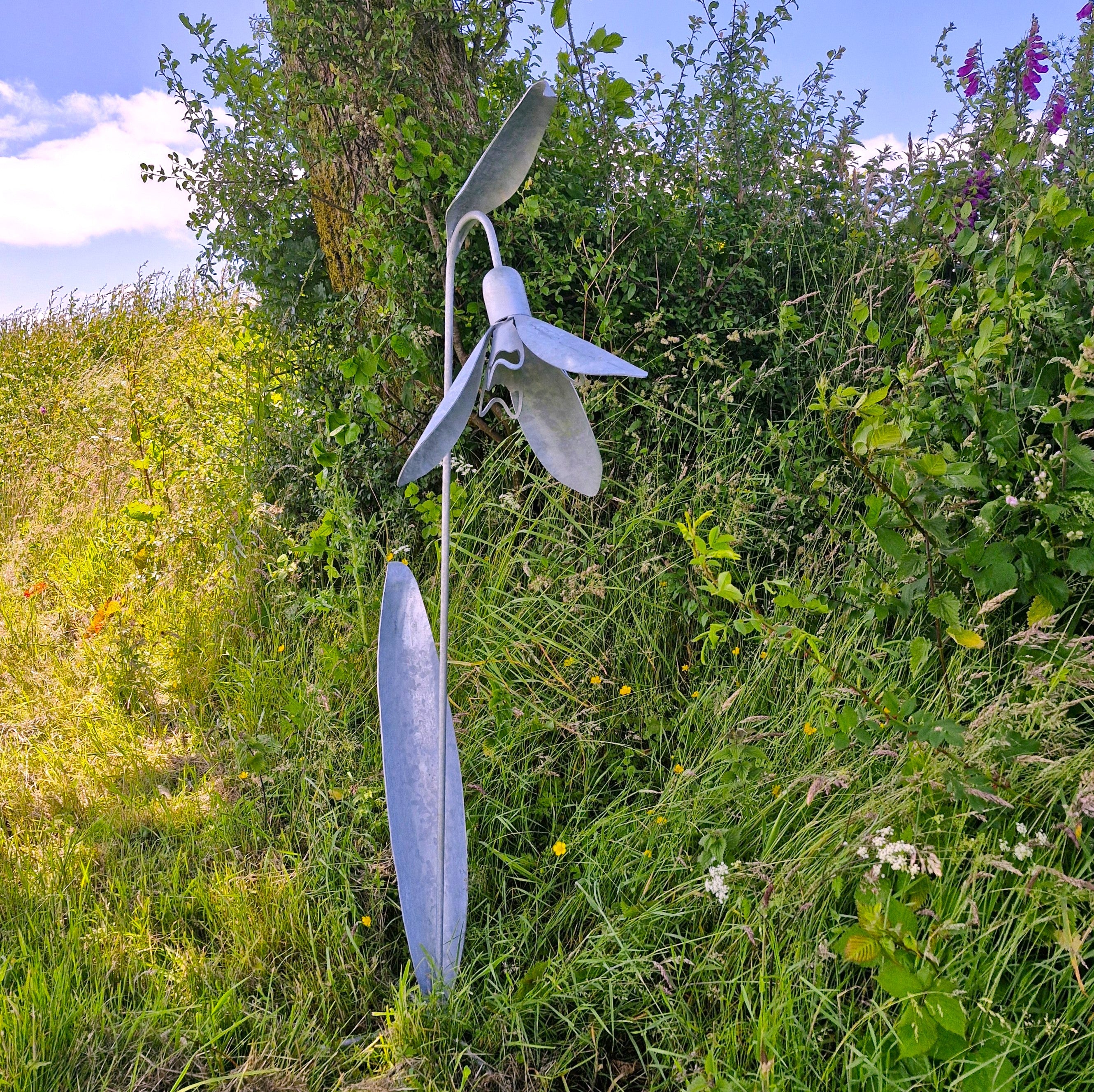 Metallic sculpture of a snowdrop in a natural setting with greenery and flowers.